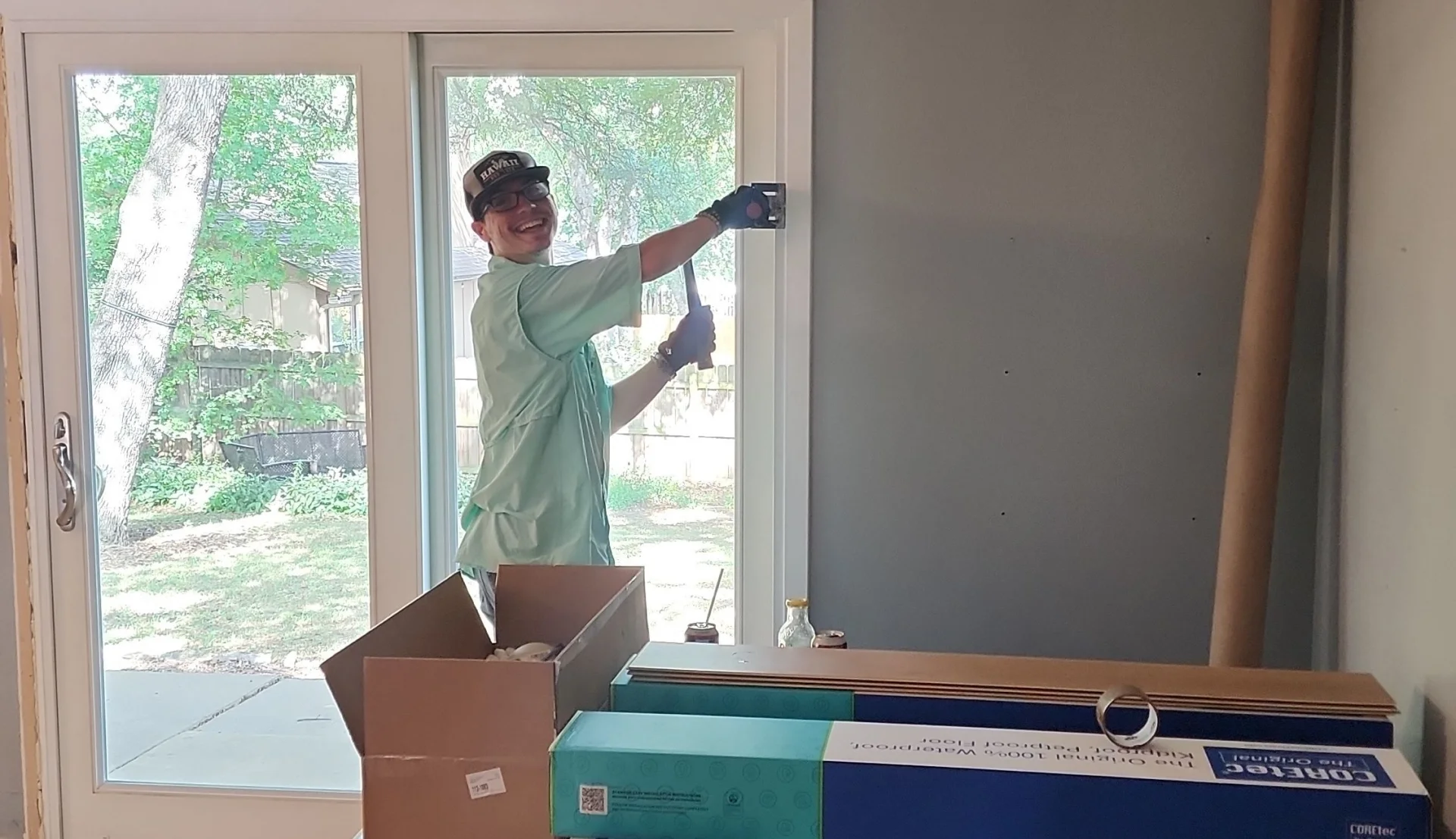 Smiling person wearing a cap, glasses, and gloves using a hammer to work on a wall next to a sliding glass door
