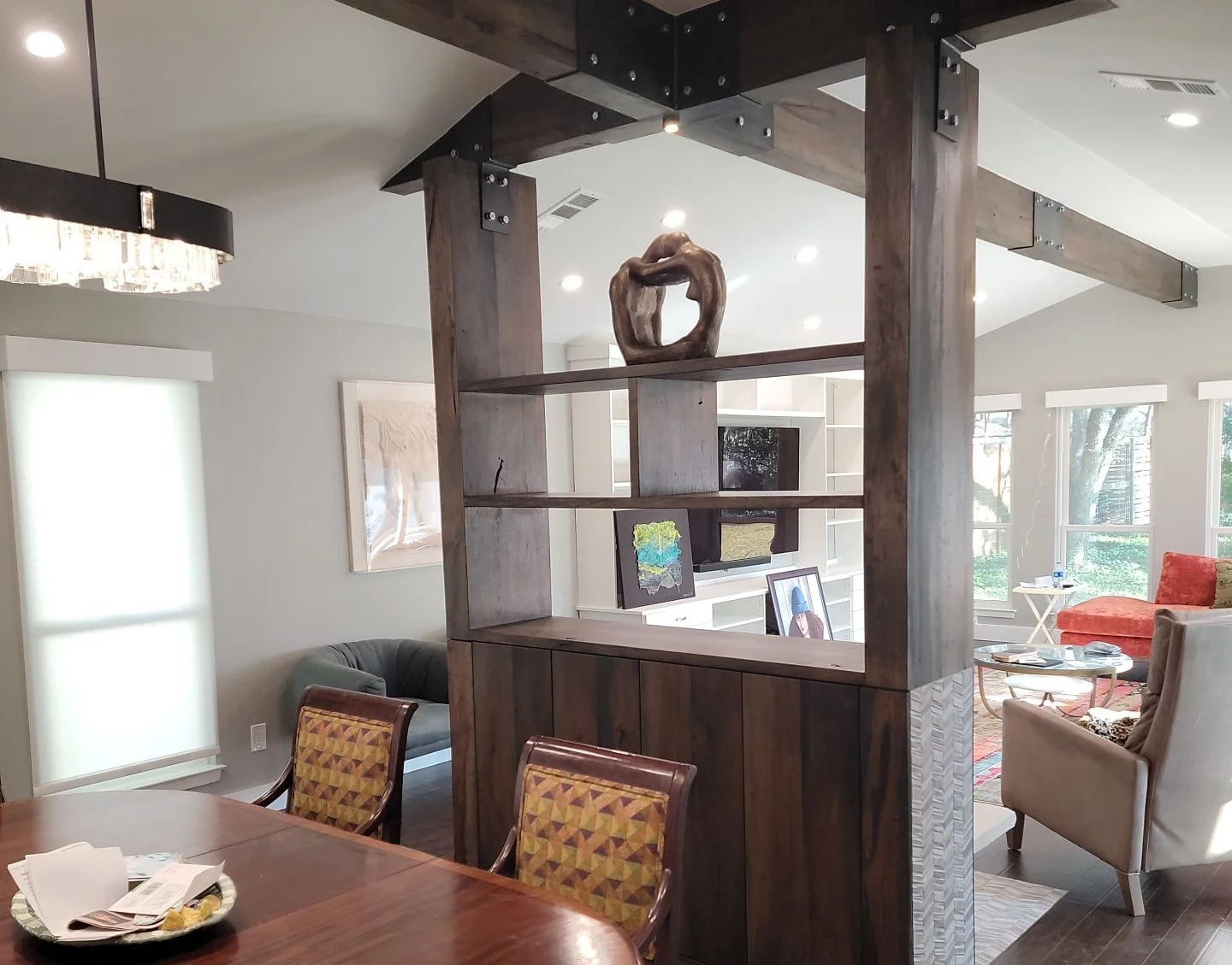 Dark wooden room divider with open shelves displaying a bronze sculpture and framed artwork, separating a dining area with patterned chairs and a living room with large windows, an orange sofa, and modern decor