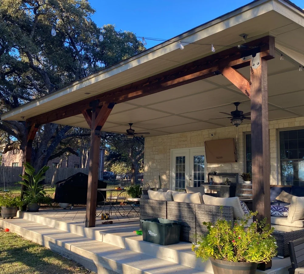 Outdoor covered patio with wooden beams and ceiling fans attached to a stone house. The patio features wicker outdoor furniture with beige cushions, potted plants, a small dining table with chairs, and string lights overhead. The area is sunlit with a clear blue sky and large trees in the background.