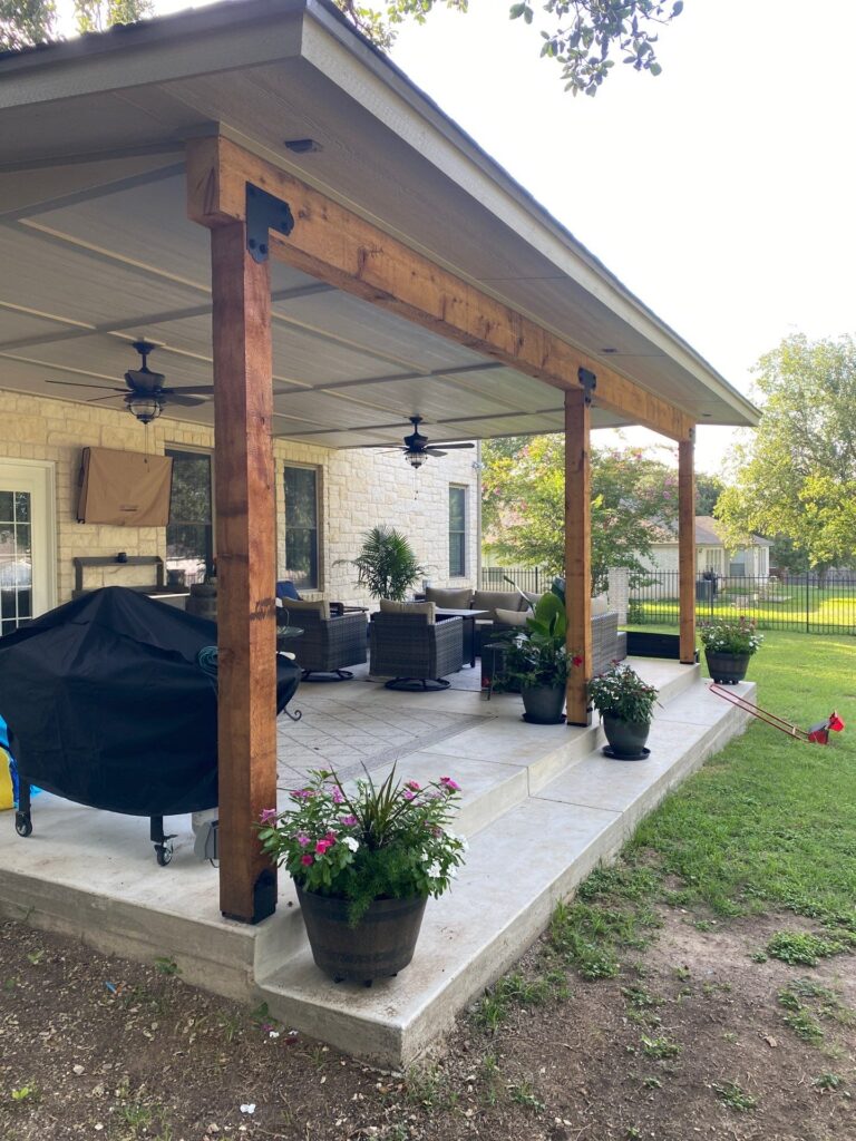 Covered outdoor patio with wooden beams, ceiling fans, wicker seating area, potted plants, and a barbecue grill, overlooking a green backyard with trees and neighboring houses.