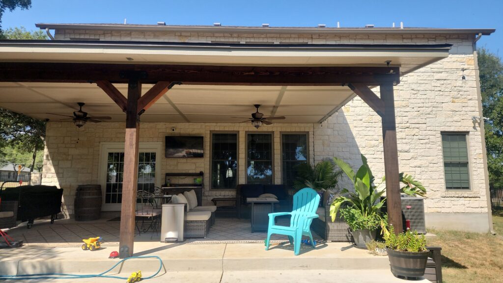 Covered outdoor patio with wooden beams and ceiling fans, featuring cushioned wicker seating, a mounted flat-screen TV, potted plants, a bright blue Adirondack chair, and children's toys scattered on the concrete floor under a clear blue sky.