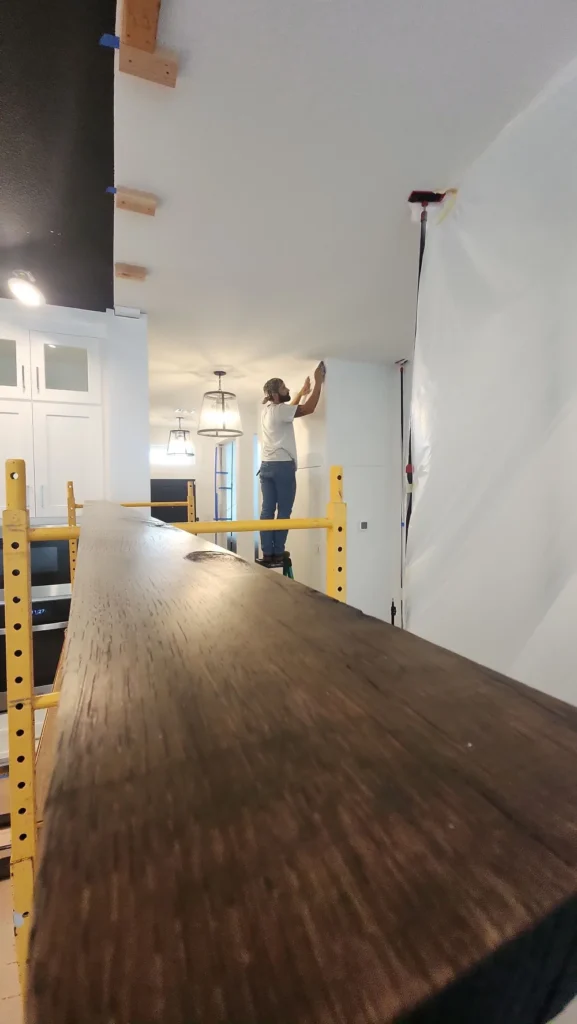 A man standing on a ladder working on a white wall near the ceiling inside a house. In the foreground, there is a long wooden beam and yellow scaffolding. The room has white cabinets and hanging light fixtures.