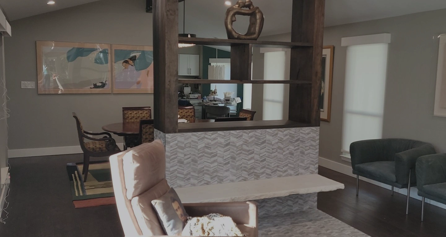Modern living room with gray walls and dark wood flooring featuring a beige recliner chair with a decorative pillow and throw blanket. The room is divided by a half wall with a geometric patterned tile and dark wooden shelving unit. In the background, there is a round dining table with upholstered chairs, framed artwork on the walls, and large windows with white blinds allowing natural light.