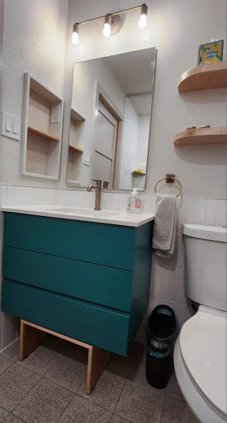Vanity with teal drawers, white countertop, modern faucet, wall mirror, towel ring, floating shelves, and toilet in a compact bathroom. Bathroom renovation project in Austin, TX.