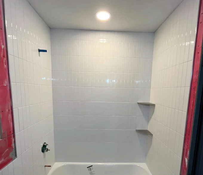 White tiled shower area with built-in corner shelves, recessed lighting, and a bathtub under construction. Bathroom renovation in Austin, TX.