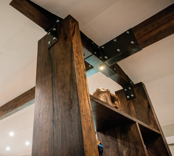 Exposed wooden beams with metal brackets supporting a ceiling, integrated with a tall wooden shelving unit in a modern interior. Dining and Living Room Renovation in Austin, TX.