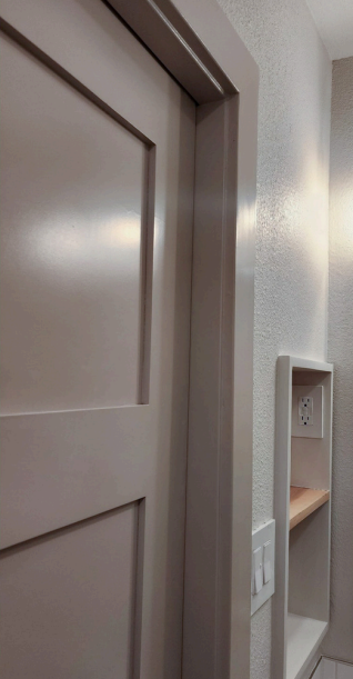 Close-up of a beige interior door with a recessed panel next to a wall niche featuring an electrical outlet and small wooden shelves. Bathroom renovation project in Austin, TX.
