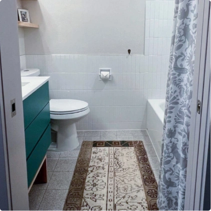 Small bathroom with white tiles, a teal vanity, a toilet, a bathtub with a patterned shower curtain, and a decorative rug. Bathroom renovation project in Austin, TX.