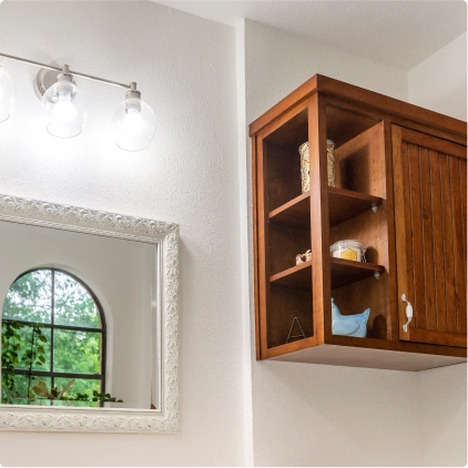 Dark stained wood wall cabinet with open shelving, three-globe vanity light, ornate framed mirror reflecting an arched window. Bathroom renovation project in Buda, TX.