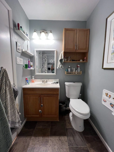Small bathroom with wooden vanity, white sink, toilet, wall-mounted cabinets, shelves with toiletries, and gray painted walls. Bathroom renovation in Austin, TX.