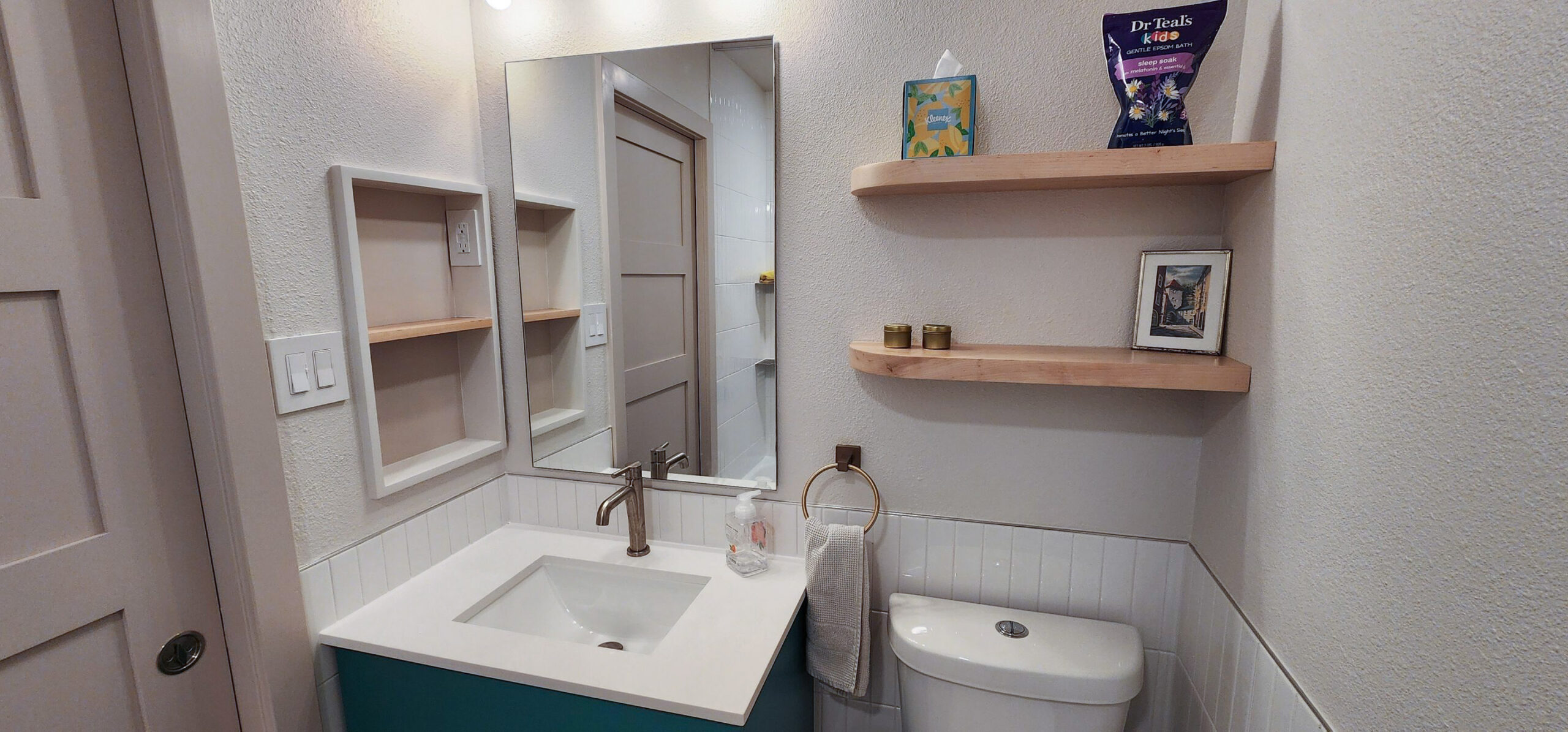 Compact bathroom with teal vanity, white countertop, rectangular mirror, towel ring, floating wooden shelves, and toilet. Bathroom renovation in Austin, TX.